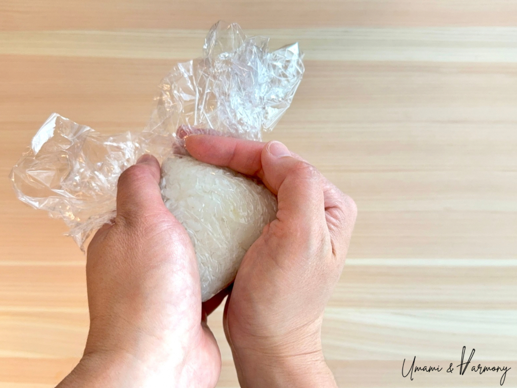 Rice being shaped into a triangle with both hands over plastic wrap.