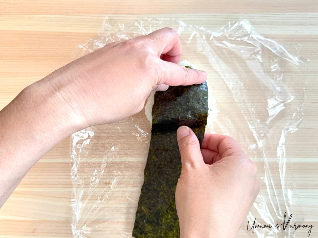 Plastic wrap removed and a nori strip being applied to the onigiri.