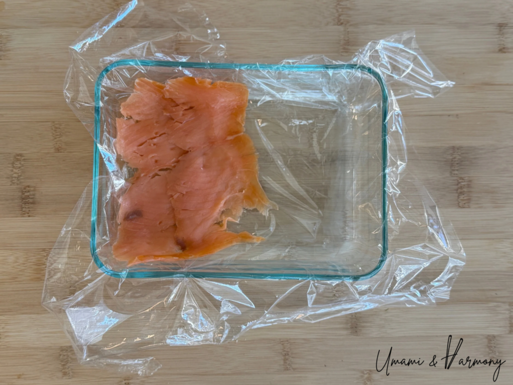 Smoked salmon slices being arranged over plastic wrap in a container