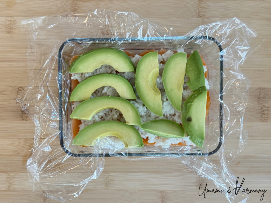 Sliced avocado being placed evenly over the rice for the second layer